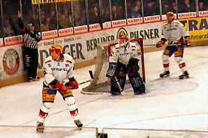 Two defencemen and a goaltender guard their goal. The referee's raised arm indicates that he intends to call a penalty.