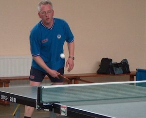 Regional competition level table tennis, showing table, net, and player getting ready to return the ball with a winning backhand topspin stroke.